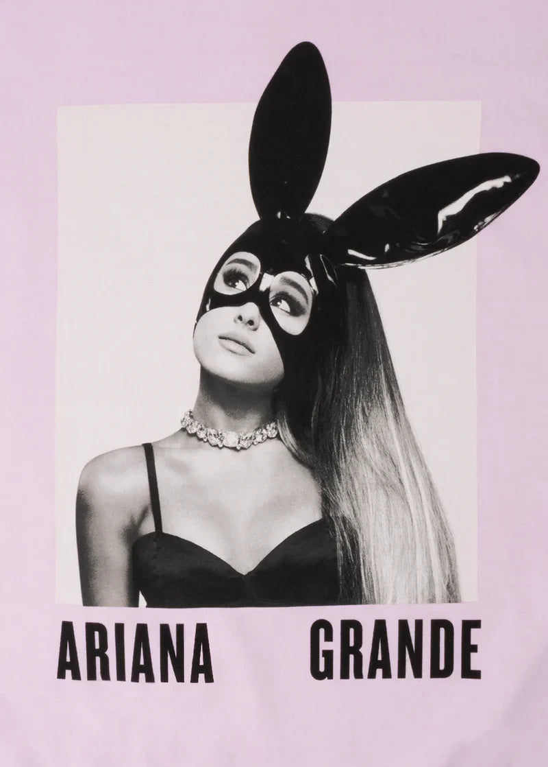 Close-up of the Dangerous Woman graphic on a pink Ariana Grande t-shirt, showing Ariana in her signature black latex bunny mask, strapless top, and crystal choker. “Ariana Grande” is printed in bold black letters below the image.