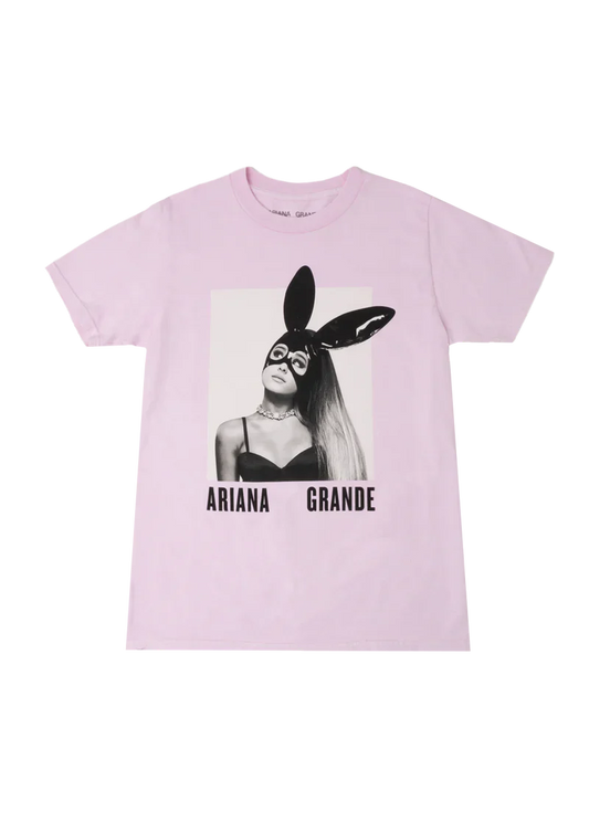 Light pink Ariana Grande t-shirt with a centered black-and-white graphic of Ariana wearing her iconic bunny mask from the Dangerous Woman album cover. The shirt features bold “Ariana Grande” text beneath the image, making it standout concert merch.