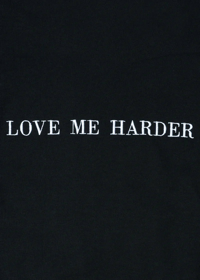 Close-up of embroidered white text “LOVE ME HARDER” on black fabric, highlighting the crisp stitch detail on Ariana Grande’s official merch.

