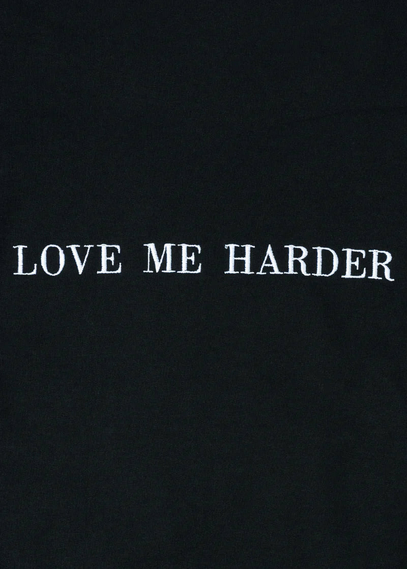 Close-up of embroidered white text “LOVE ME HARDER” on black fabric, highlighting the crisp stitch detail on Ariana Grande’s official merch.

