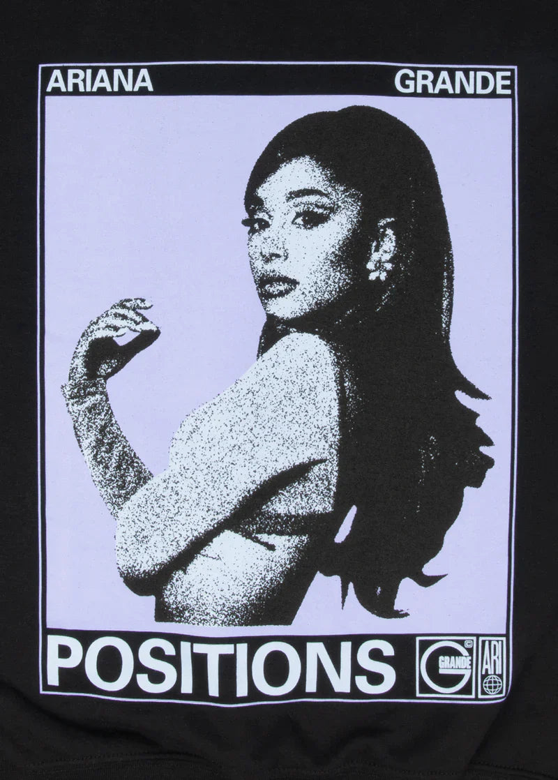 Close-up of the “Positions” crewneck graphic showing Ariana Grande in a grainy black-and-white halftone effect with lavender background. The design is framed with “Ariana Grande” at the top and “POSITIONS” in bold white below.

